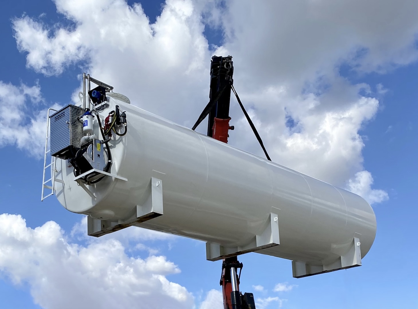 White cylindrical fuel storage tank being lifted by crane against a blue sky