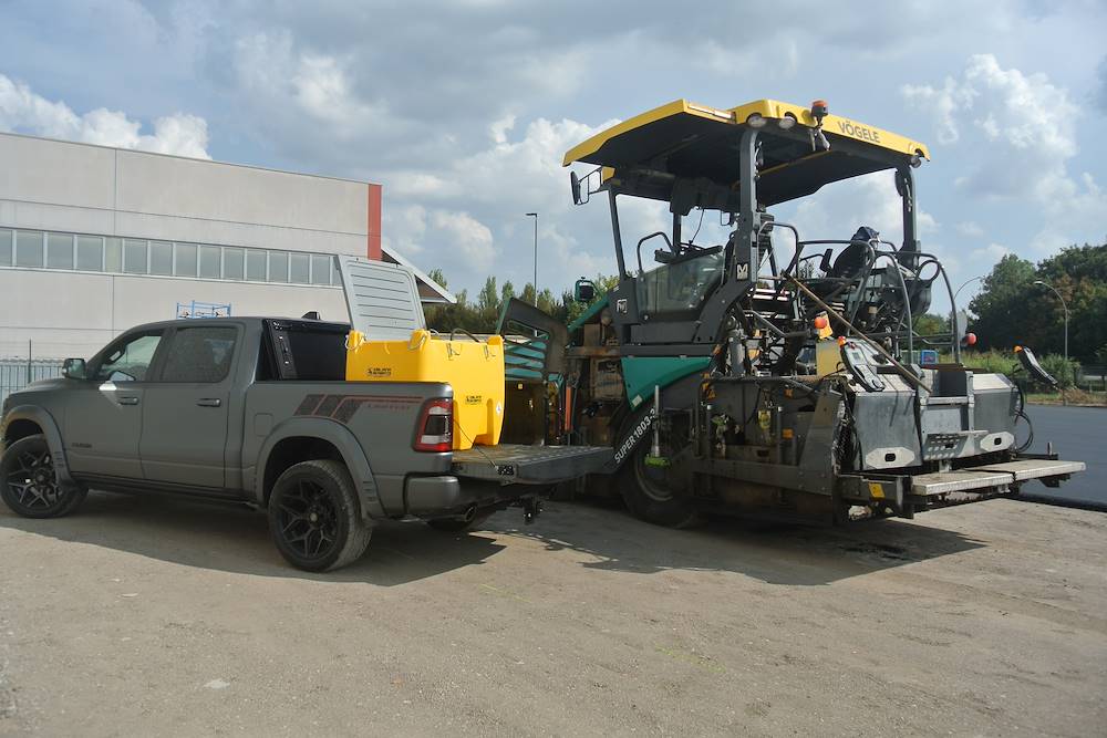 Pickup truck with a fuel tank parked beside a road paver on a construction site