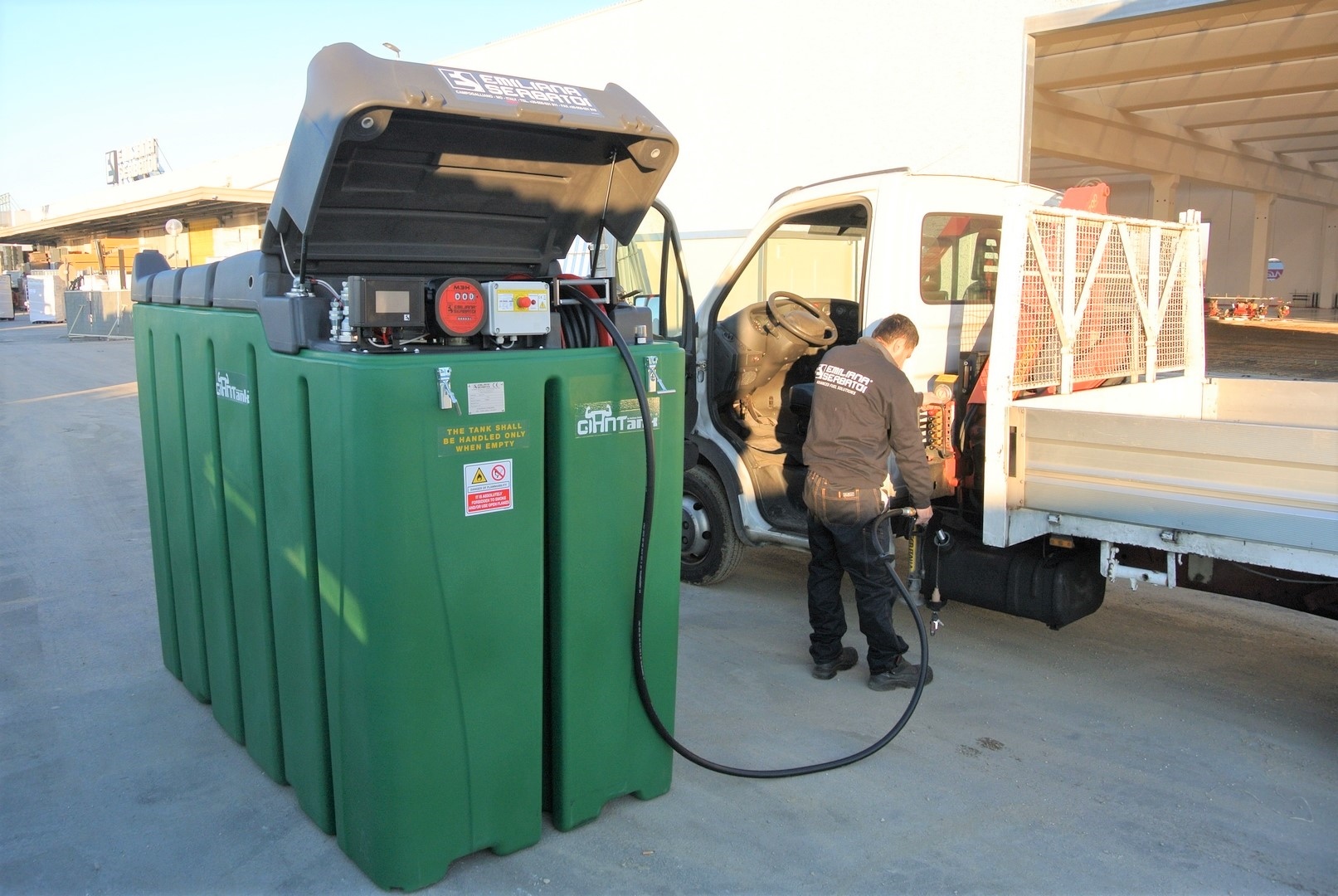 Man refueling a flatbed truck from a green vertical storage tank at an industrial yard